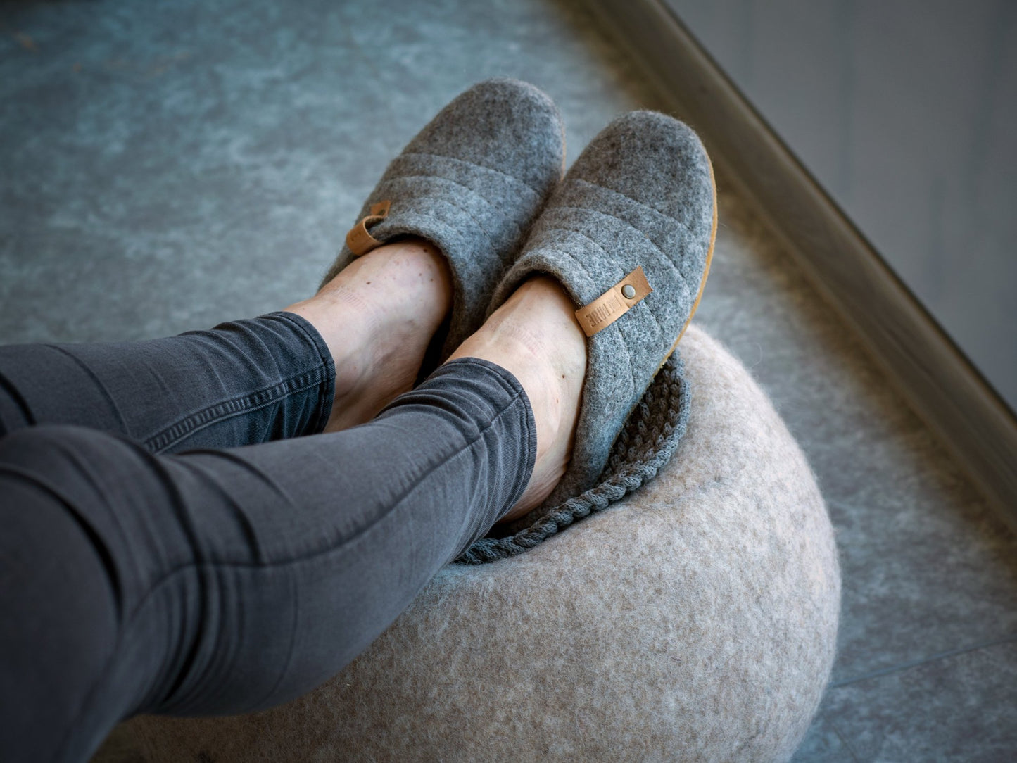 A woman wearing Cocoon felted wool slippers with collapsible backs, with her legs on a felted wool ball.